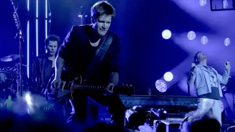 A guitarist with black clothing on stage with a band and two other members of the band.