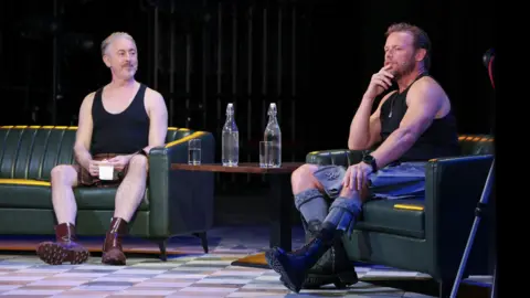 PA Media Two speakers sit on green leather armchairs on a stage during a live discussion. A small table between them holds two glass bottles of water and two drinking glasses. Both speakers wear black sleeveless tops with kilt-style skirts and boots, and one speaker is wearing a headset microphone while speaking. The stage is lit with blue and purple lighting against a dark background.