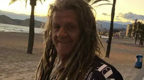 A smiling man with long dreadlocks. He is wearing a black t-shirt with white writing. He is stood on a beach and in the background is the sea and sand. 