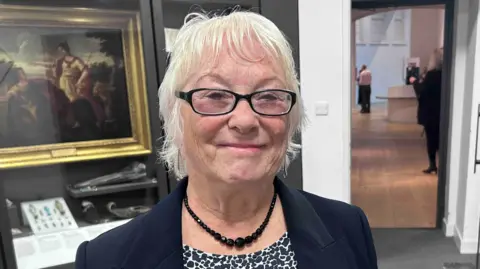 Kate Bradbrook/BBC Danielle Stone is looking towards the camera smiling. She has short white hair and is wearing a black and white T-shirt, a black blazer and a black necklace. Behind her is a painting and a door leading to another room.