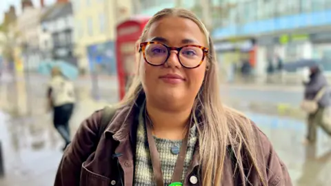 DAVE EDWARDS/BBC A young woman with glasses looks at the camera. Behind her is a busy shopping street on a winter's day.