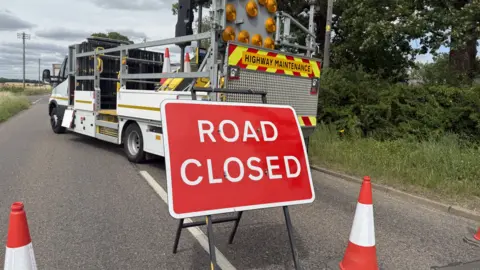 Elliot Deady/BBC A road has been closed and has cones going across it. There is a sign in the middle of the road which is red and has ROAD CLOSED on it in white writing. A truck is parked across the road.