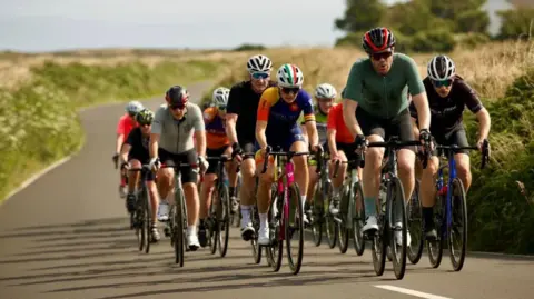 Gran Fondo Isle of Man A group of people on bicycles wearing multicoloured jerseys and helmets cycle on a road in the countryside