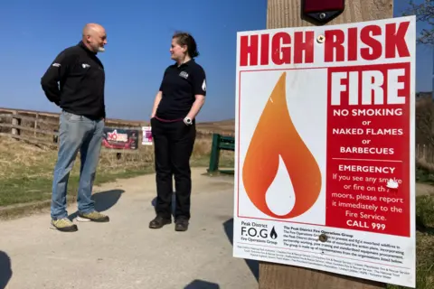 A poster on a wooden fence warning about HIGH RISK FIRE - detailing the warnings around wildfires in the Peak District. A man and a woman are stood in the background.