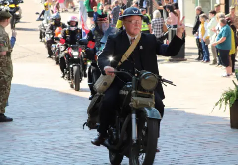 Callum Malone an older man riding a motor cycle while holding is left hand in a v-shape. He is wearing old fashioned clothes.