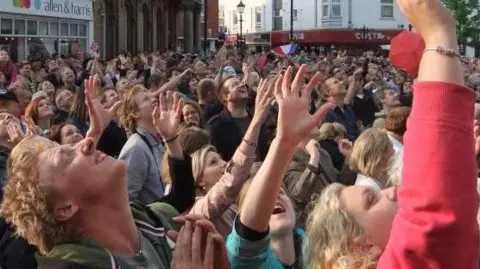 Abingdon Town Council A huge crowd of people in a town centre look up towards the sky and reach their arms out expectantly. 