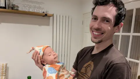 A man in a brown t-shirt smiles at the camera while holding his baby son