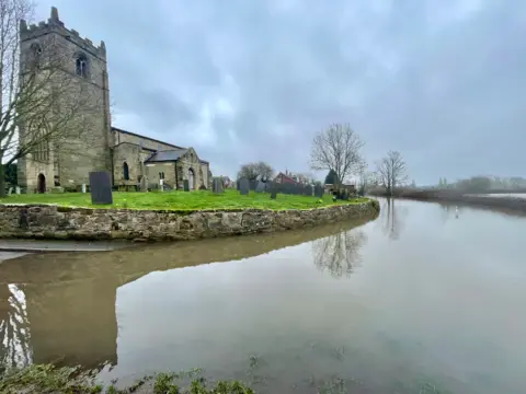 An old stone church stands on a grassy graveyard with a stone wall dropping down to the corner of a road. The road is completely flooded in brown water. The hedgerows alongside the other side of the road poke out of the water, which continues into the fields behind. The sky is cloudy.