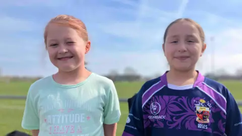 Anna and Maisie Jane, with one wearing a turquoise attitude t shirt and the other in a Swindon Roses top.
They both have their hair up and are smiling at the camera. There is a rugby pitch behind them which is out of focus.
