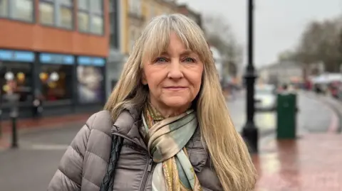Cllr Karen Rowland is wearing a grey puffer coat, an autumnal coloured scarf, with blonde hair. A Reading Street is in the background.