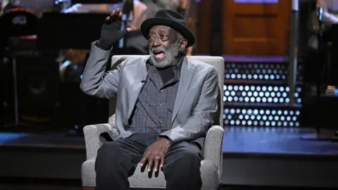 Todd Owyoung/NBC via Getty Images Garrett Morris sits on a light grey armchair holding a hand in a fingerless glove up. He wears a black hat, grey jacket and black trousers