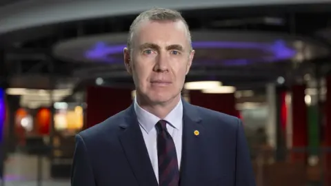 Adam Price looking at the camera. He is standing in front of desks in the BBC office. 