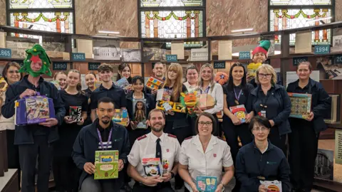 A group of men and women posing for a photo, some wear navy shirts and jumpers, while others wear white salvation army shirts, each hold a children's toy.