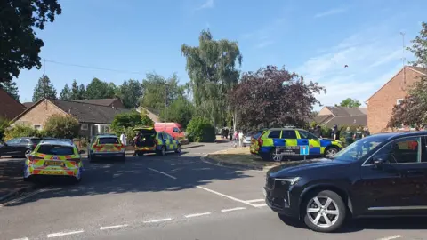 Four police cars at the entrance to a residential road. A black car is turning into the road. Officers can be seen clustered outside a red brick house in the background.