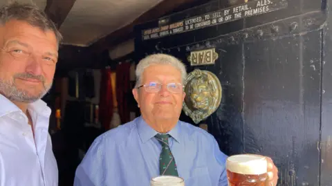A man with grey hair and glasses is holding a cup of beer in front of a door. The door has an upside-down sign that reads "bar". There is a large brass lion face on the door. Next to the man is another man with dark hair and grey facial hair