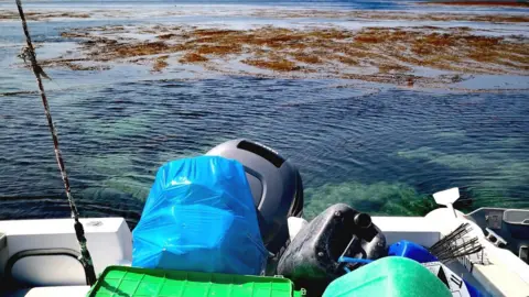 Nikki Banfield/ BareFoot Photographer Litter bagged up on a boat. The boat is on water which is blue and clear. The boat is white and there is seaweed on the surface of the water.