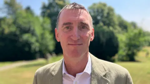 A man in a beige suit jacket stood in front of some trees.