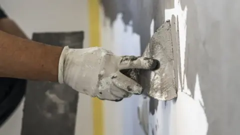 A person is spreading grey plater on a white wall. The person is wearing a white glove. There is yellow tape on the corner of the wall. 