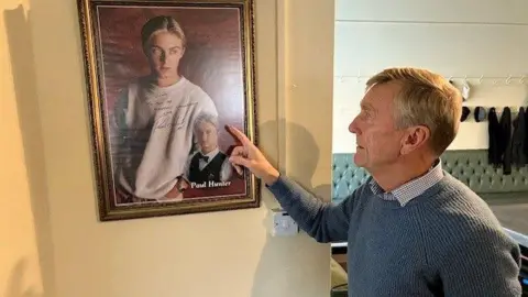 David Spereall/BBC A man in a blue jumper points to a signed photo of Paul Hunter on the wall.