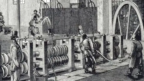 Universal History Archive/Universal Images Group/Getty Images A black and white illustration of iron being produced in Dowlais in the 19th Century. A man in a top hat and suit watches men at work wearing protective visors standing by a long piece of machinery.