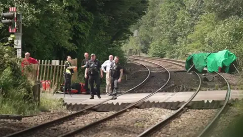 The railway tracks at the crossing showing emergency service people to the left and a green tarpaulin to the right covering up damage