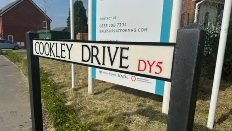 Close-up of a road sign saying Cookley Drive with DY5 in red letting, in front of grass, a housing development sign, and new houses.