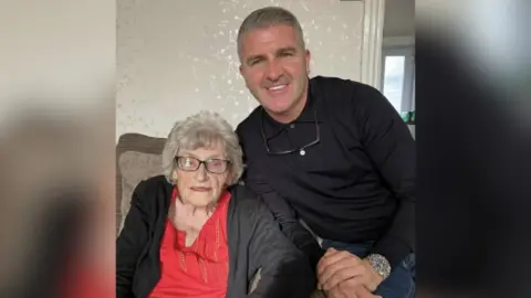 Hannah Kent Two people are sitting together on a beige sofa in a living room. Older woman, Joan is wearing a bright red top under a black cardigan, while the other, middle-aged man with silver hair, Ryan, is dressed in a dark long-sleeved shirt and jeans, leaning slightly forward with hands clasped. A pair of glasses hangs from the neckline of the second person’s shirt, and a silver watch is visible on the wrist. The wallpaper behind them has a subtle floral pattern.