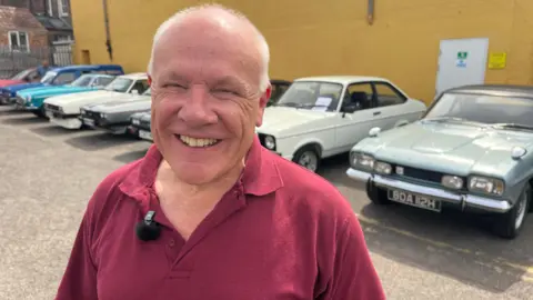 A man smiles widely while stood in front of classic cars which are parked in a row in front of a yellow wall. He has short get hair and is wearing a burgundy polo shirt.