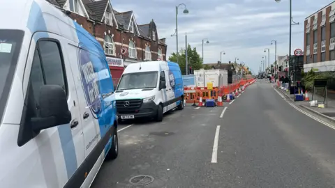 Two Southern Water vans on the road with traffic cones stretching down the centre of a two way road into the distance. On the left a digger and shipping container are visible.
