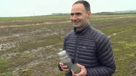Jim Mann standing in a field holding a jar of the basalt rock powder. He has short dark hair and is wearing a black puffer jacket.