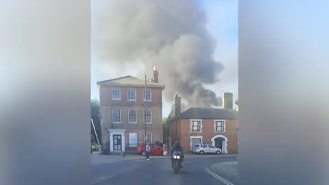 A large smoke plume can be seen billowing in the area behind several buildings. A motorcyclist can be seen on a road travelling toward the camera.