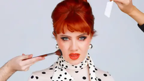 The picture shows a person with bright red hair styled in an up‑do, wearing a cream top with black polka dots and a matching scarf tied at the neck. Two people positioned just out of frame are working on them, one holding a makeup brush near the jawline and the other holding what looks like a tissue or blotting paper. The scene looks like a behind‑the‑scenes moment during a makeup or styling session.