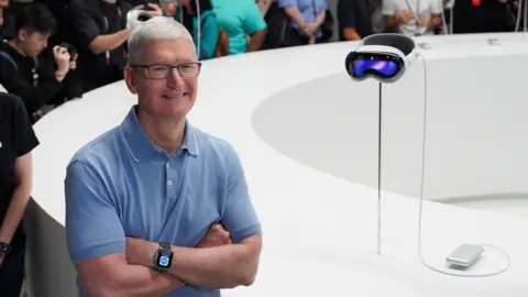 Reuters Tim Cook wearing a light blue polo shirt, standing next to the Apple Vision Pro headset.