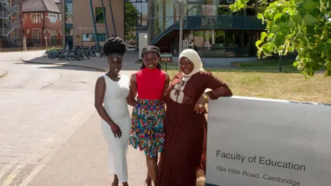 University of Cambridge Francisca Arhinful, Jemimah Mensah, Fadila Issah, lined up together and smiling broadly. They are standing outside Cambridge University's Faculty of Education, beside its sign. Behind them is grass, a road and a brick and glass building with many bicycles in racks. 