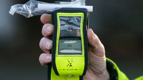 PA Media Close up of a police officer's hand holding a fluorescent yellow breathalyser. The sleeve of the high visibility jacket is also fluorescent yellow. The mouthpiece is covered in plastic wrapping. 