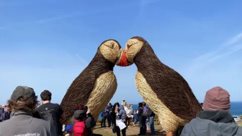 A wide, side shot of the two giant puffin sculptures with their heads together and crowds all around. The willow is not frayed as this was taken before the storm. A clear blue sky is visible in the background.