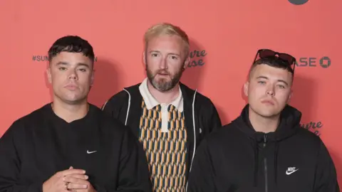 Three men all wearing black sweaters are stood side by side looking directly at the camera. They have serious expressions. 