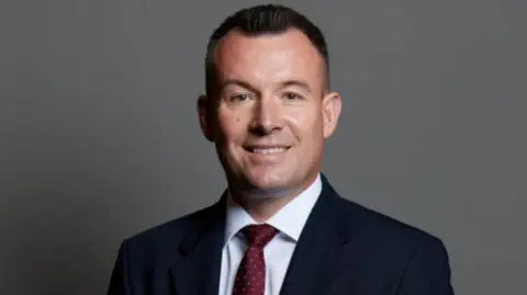 A man with short dark hair, wearing a dark suit, white shirt and red tie against a grey background