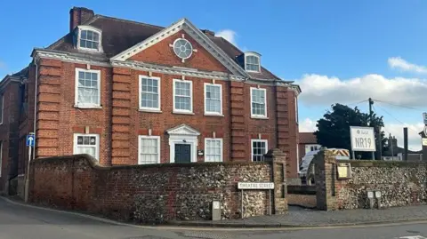 Aimee Dexter/BBC A bricked building is on the left. It is three storeys, and has white windows and a black door in the middle. A cobblestone wall goes around the front of the building which is on a road. 