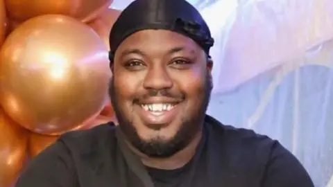 A smiling man stands facing the camera with arms crossed, wearing a black T‑shirt and a black head covering. Gold balloons and a light backdrop are behind them.