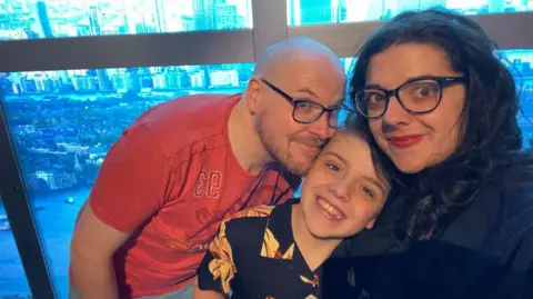 Three people ride in the London Eye. A man with a bald head,wearing black framed spectacles and a red T-shirt has his face against the face of a young boy in a black T-shirt who is leaning against a woman with black hair and black spectacles.