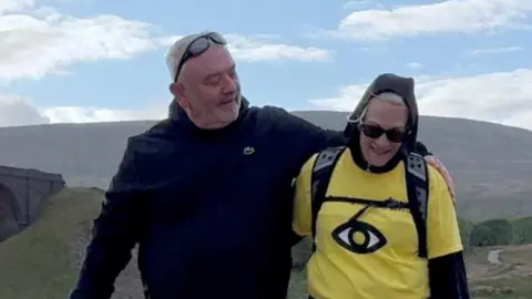 Contributed Mrs Blezard walks with her arm around her husband's waist. He is looking at her as they walk with his arm also around her shoulders. He has short grey hair and has sunglasses on his head.