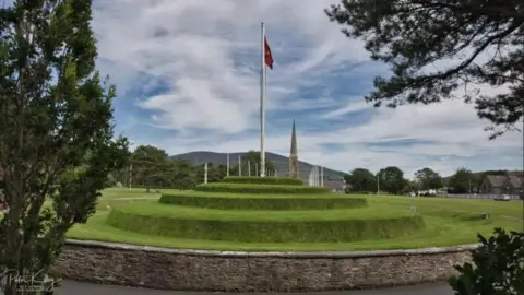 MANX SCENES Tynwald Hill, which has round grass tiers with a flagpole on top. It is surrounded by a Manx stone wall and there are trees nearby,