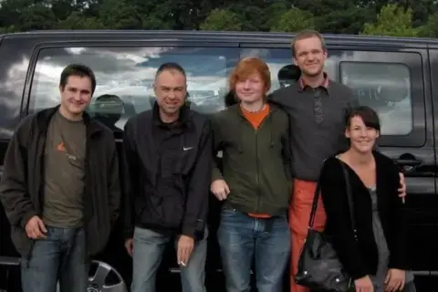 John Parker Ed Sheeran, centre, wears an orange t-shirt and green hoodie with jeans. To his left Luke Concannon from Nizlopi wears a grey jumper and orange trousers. He has his arm around a woman and there are two other unnamed men to Sheeran's right.
