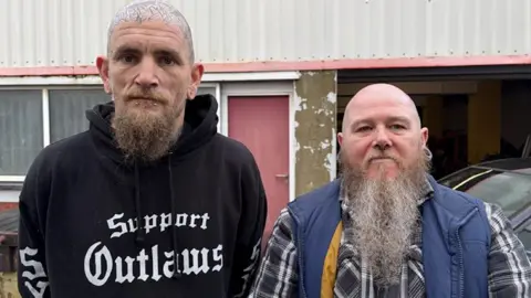 Wesley and Marc pictured standing next to each other in front of a metal warehouse unit. They are both looking at the camera with a serious look. Wesley has a bald headed that is tattooed and a brown beard. He has a black hoody on. Marc has a blue gilet on with a tartan shirt under it. He is also bald and has a longer grey beard.