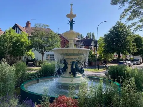 BBC / Sally Fairfax An ornate fountain surrounded by flowers and plants, gushing water in the middle of tree-lined, residential street on a sunny day