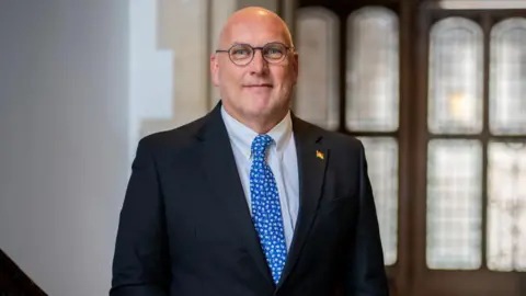 Hampshire County Council leader Nick Adams-King posing for the camera. He is wearing round glasses and a dark suit.