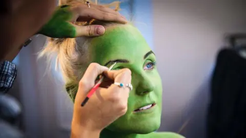 Kerry Ellis Ms Ellis sits in a chair as a make up artist paints her eyebrows. She has green face paint on and she is looking away from the camera. 