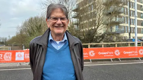 Sir John Madejski smiling at the camera with is arms behind his back. He is wearing a blue shirt, with a v-neck blue jumper on top and a brown leather jacket.