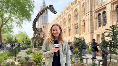 BBC Journalist Sophia Herod, smiling with a microphone outside London’s Natural History museum on a sunny day with trees and a dinosaur sculpture behind her